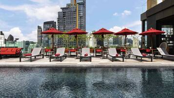 Un espace piscine aménagé sur le toit, avec des chaises longues et des parasols rouges, entouré d’immeubles modernes.