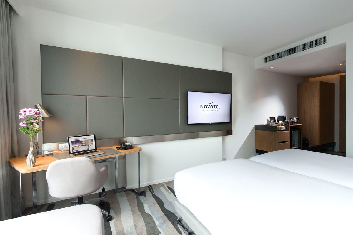 Habitación de hotel con cama, escritorio con computadora portátil, televisor montado en la pared y una pequeña mesa con un jarrón de flores.