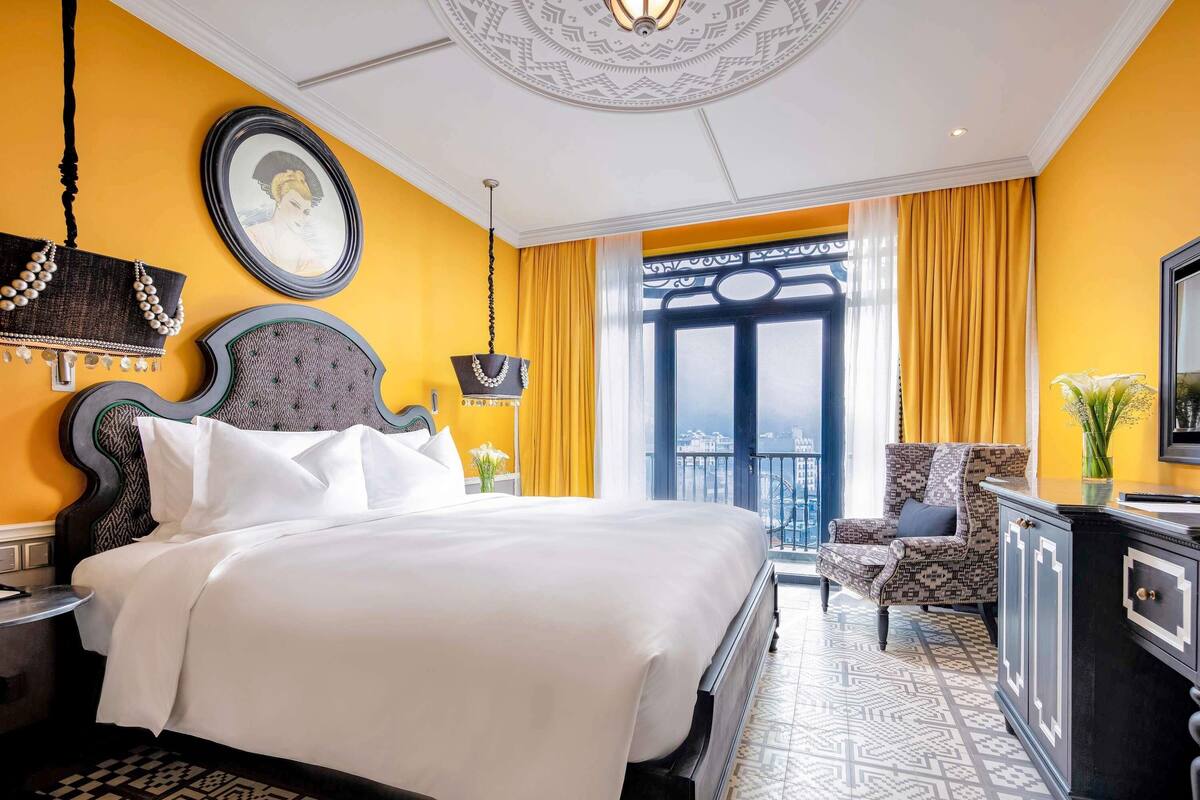 A hotel room with a large bed, a desk, a chair, a view of the city through a balcony, and a patterned floor.