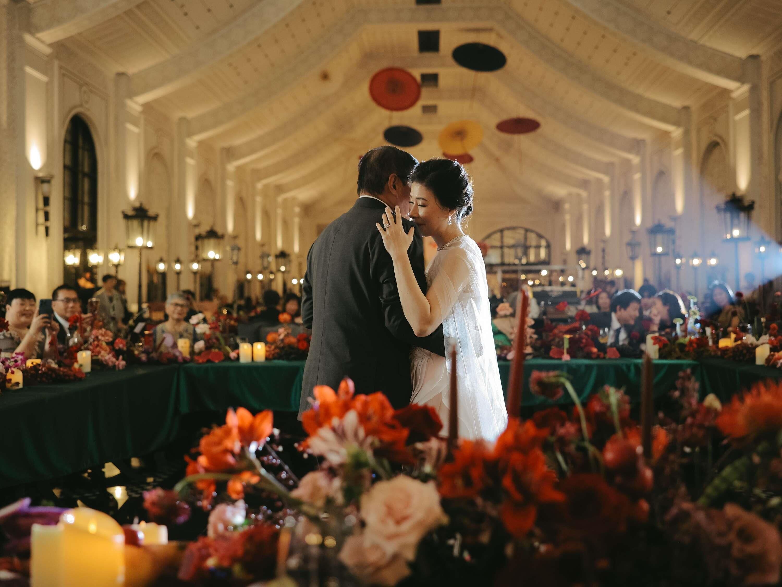 A couple kissing at a wedding reception in a hall with high ceilings and chandeliers.