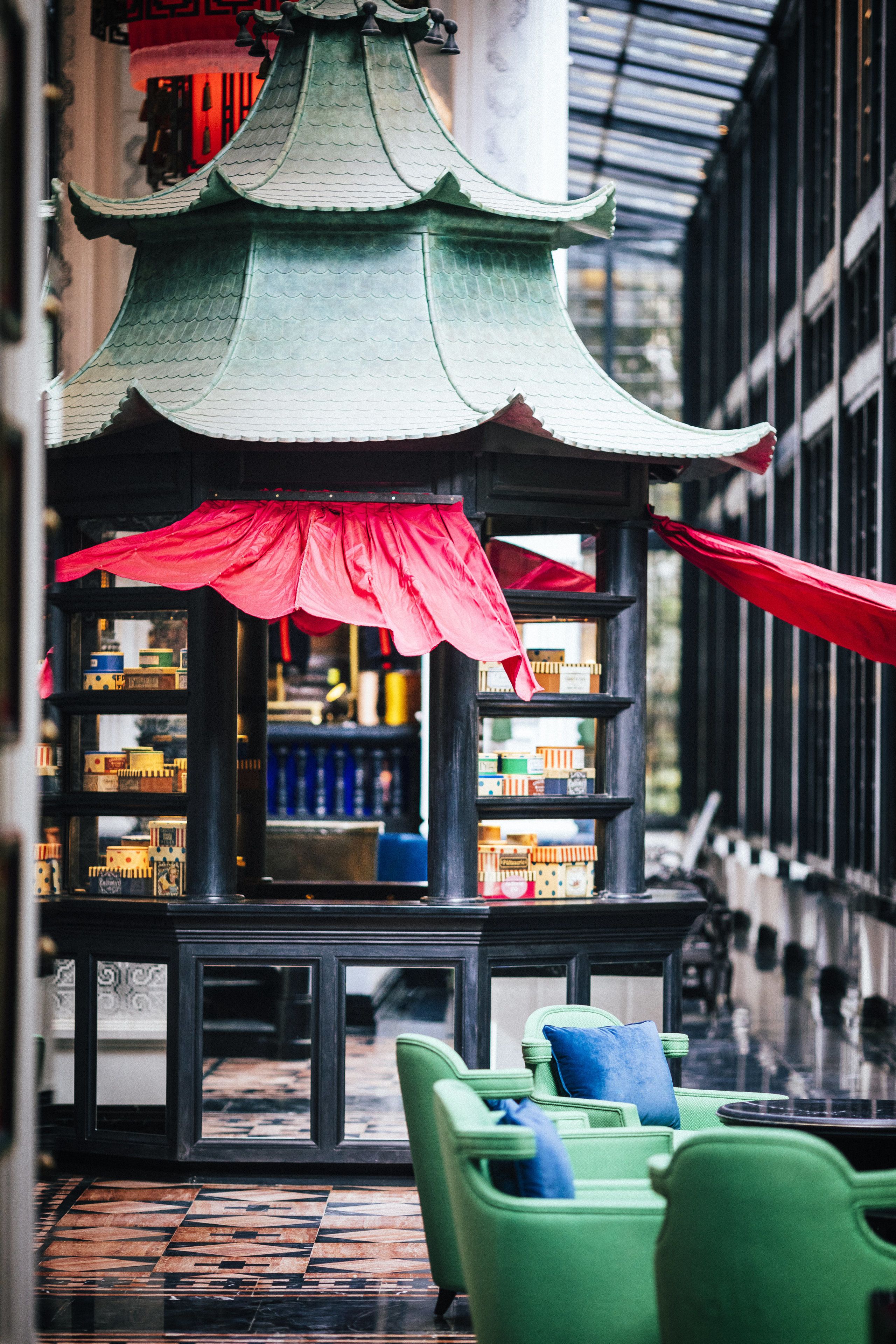 Un pavillon de style asiatique traditionnel, doté d’un toit en tuiles et d’une marquise rouge, entouré de chaises vertes et d’étagères contenant divers objets.