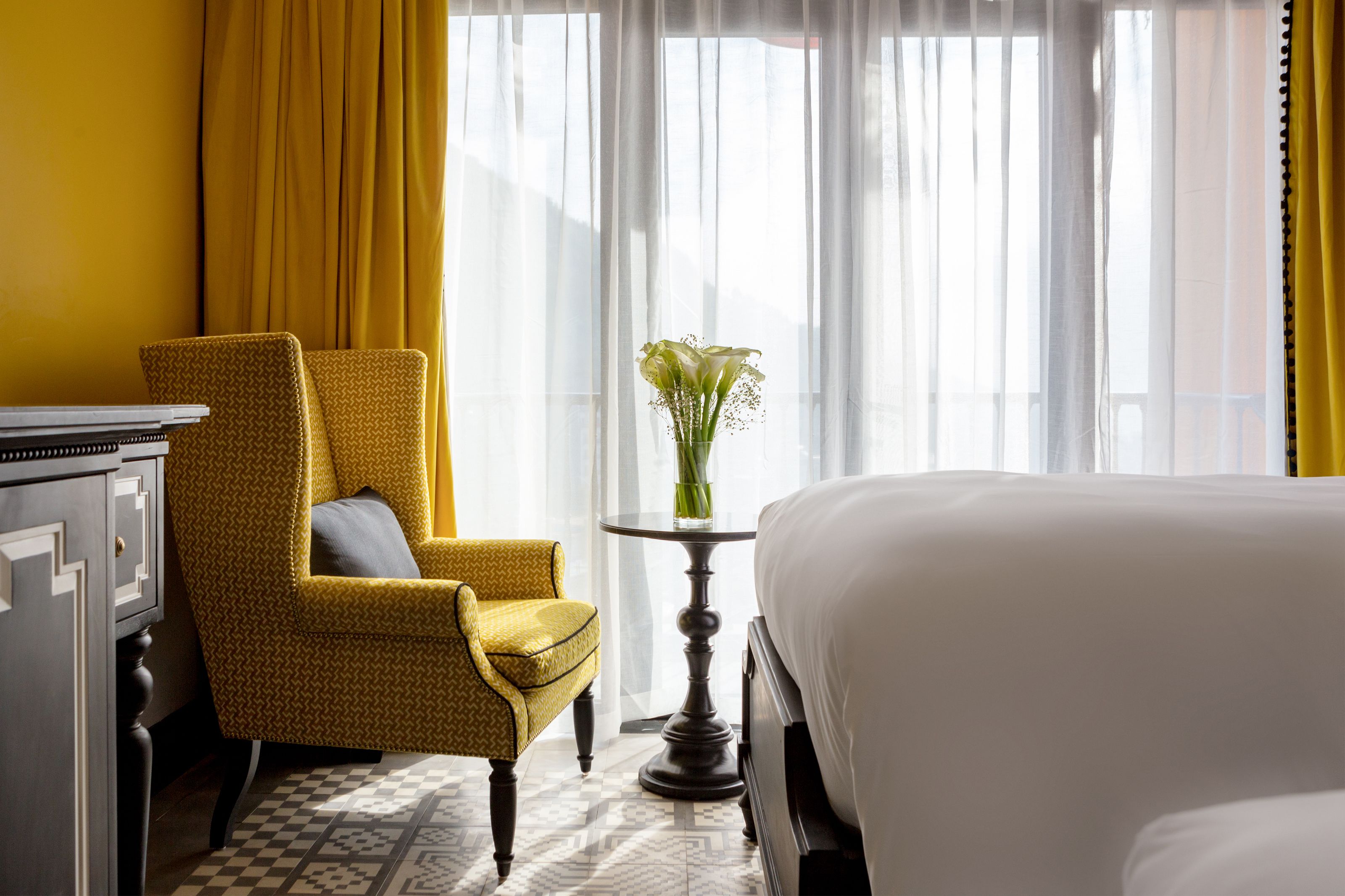 Habitación de hotel con una cama, un sillón amarillo, una mesita con un jarrón de flores y una ventana con cortinas de encaje.