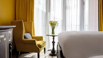 Habitación de hotel con una cama, un sillón amarillo, una mesita con un jarrón de flores y una ventana con cortinas de encaje.