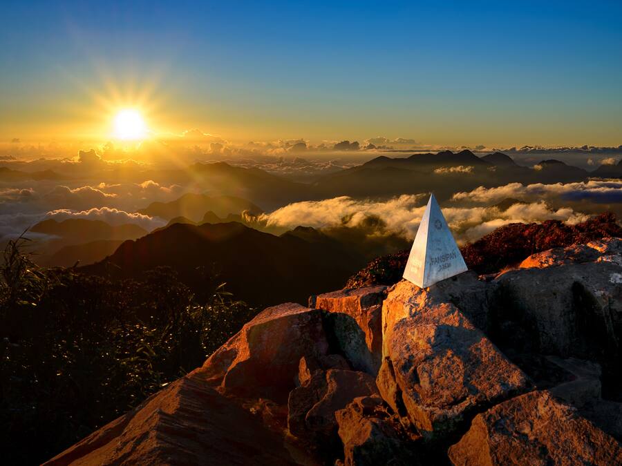 A mountain peak with a flag, surrounded by clouds and mountains under a clear sky.