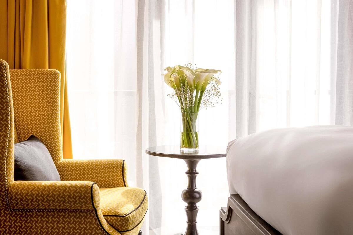 A hotel room with a bed, a chair, a small table with a vase of flowers, and sheer curtains.