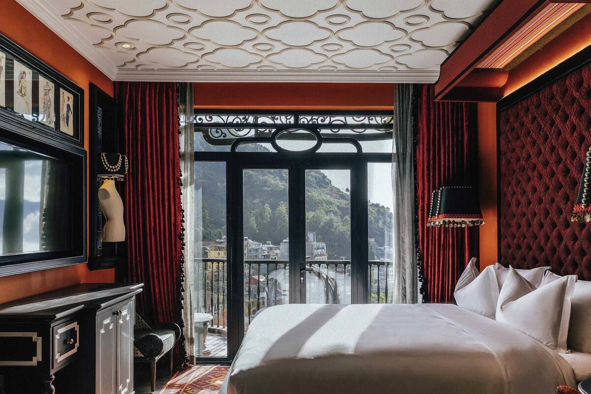 A hotel room with a large bed, a TV, a dresser, and a balcony with a view of buildings and hills.
