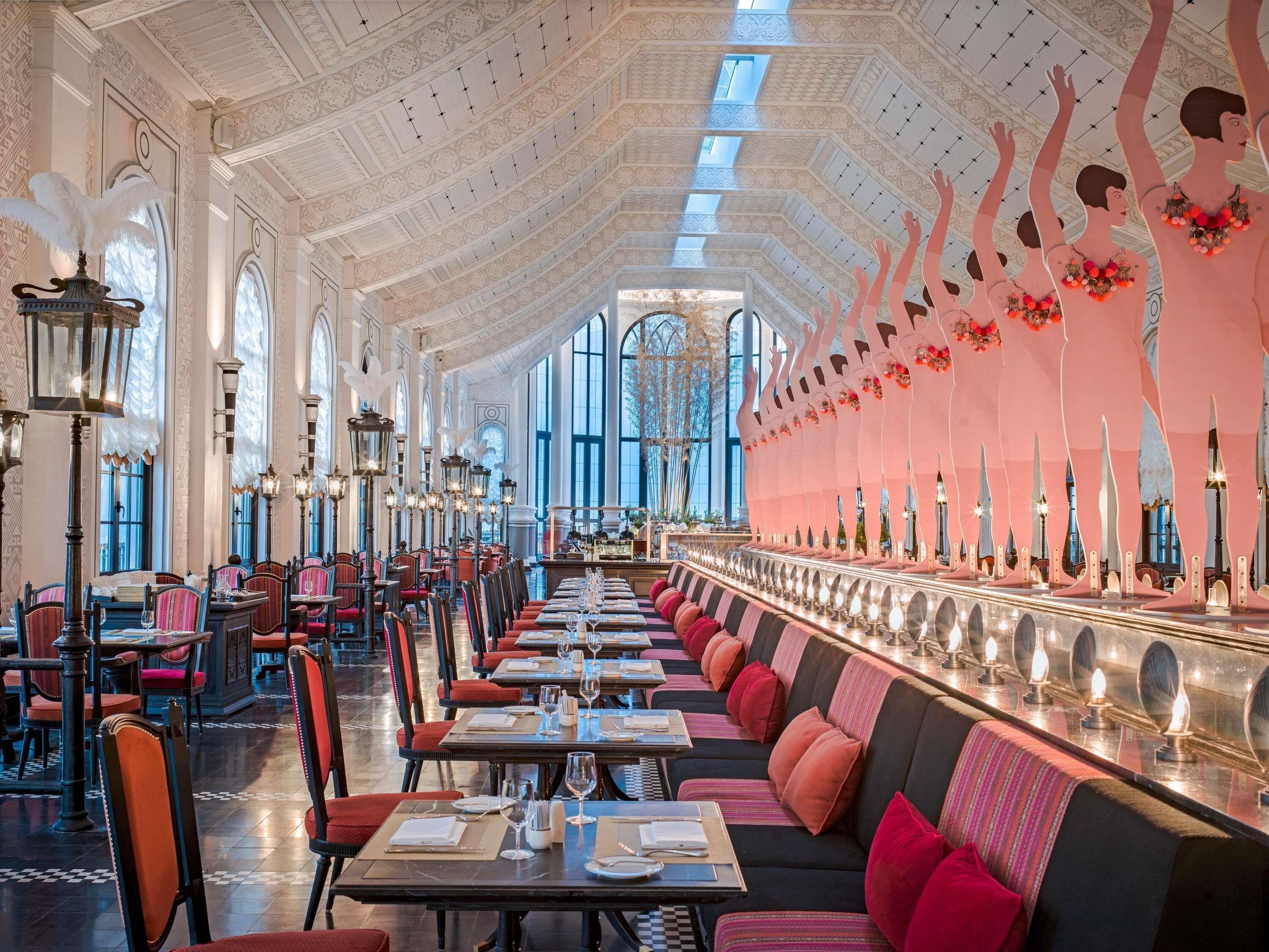 A spacious, elegantly decorated restaurant with high ceilings, ornate chandeliers, and a long dining area with plush seating.
