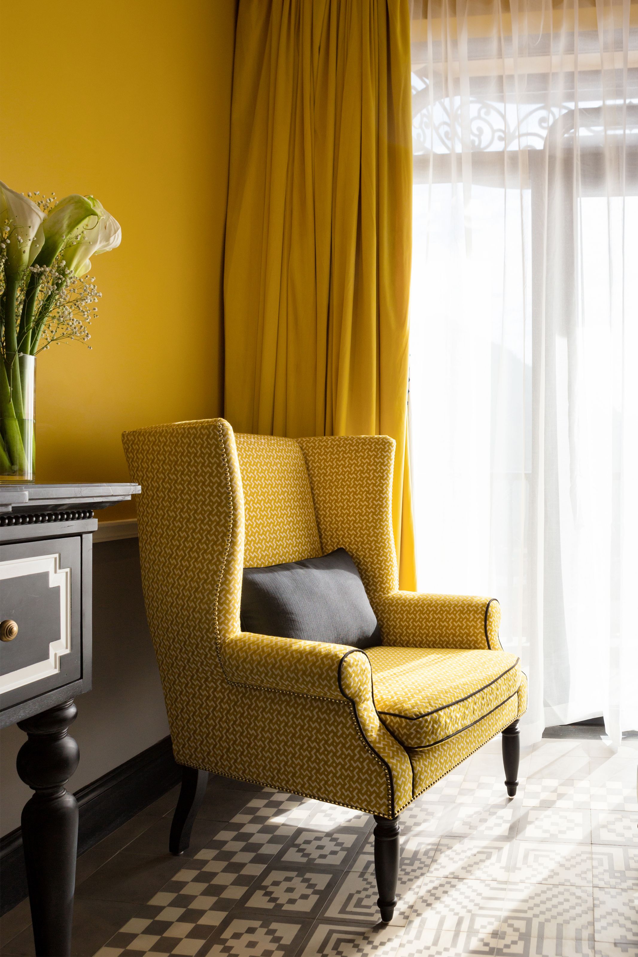 A yellow armchair with a patterned cushion, a vase with flowers, and a patterned floor.