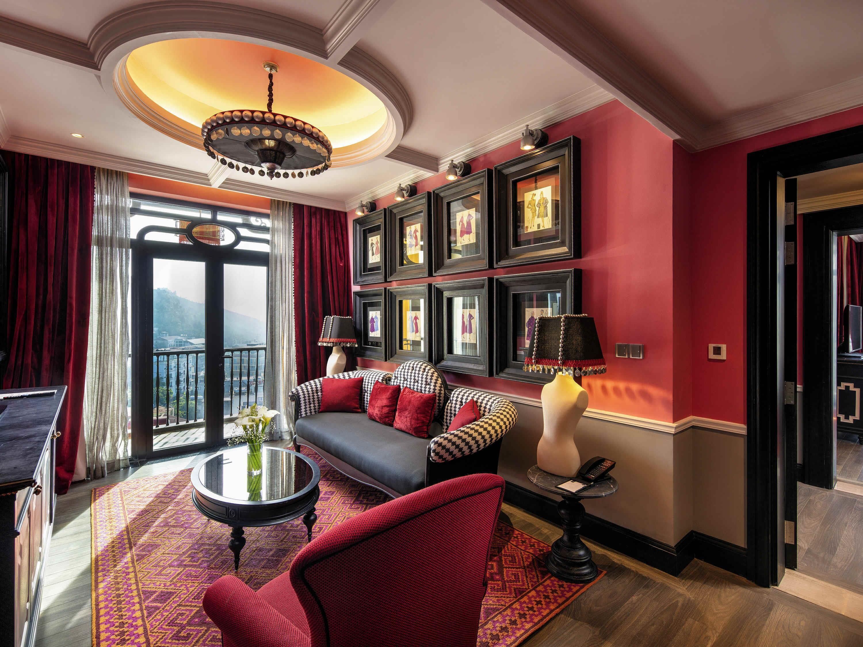 A room with a sofa, armchairs, a round coffee table, a chandelier, framed artwork on the wall, and a balcony view.