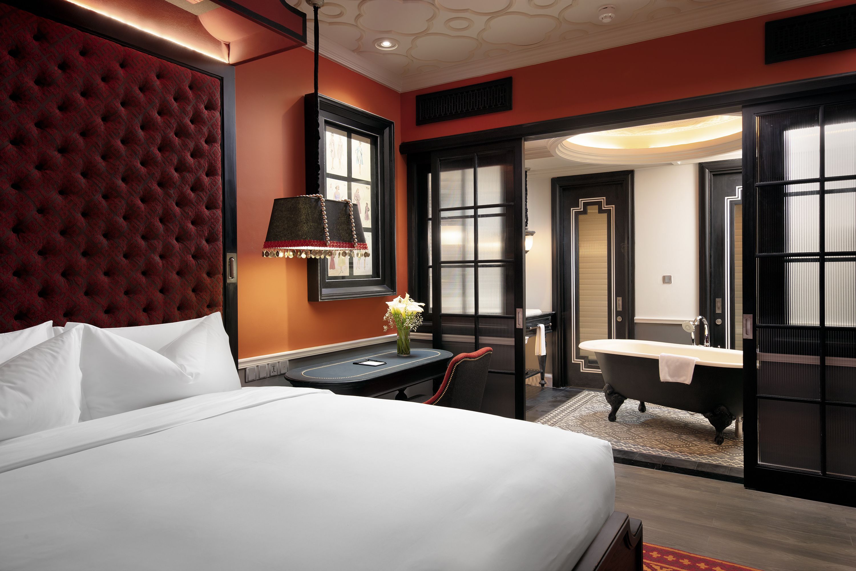 A hotel room with a large bed, a desk with a lamp, a chair, a bathtub, and a bathroom with a mirror and sink.