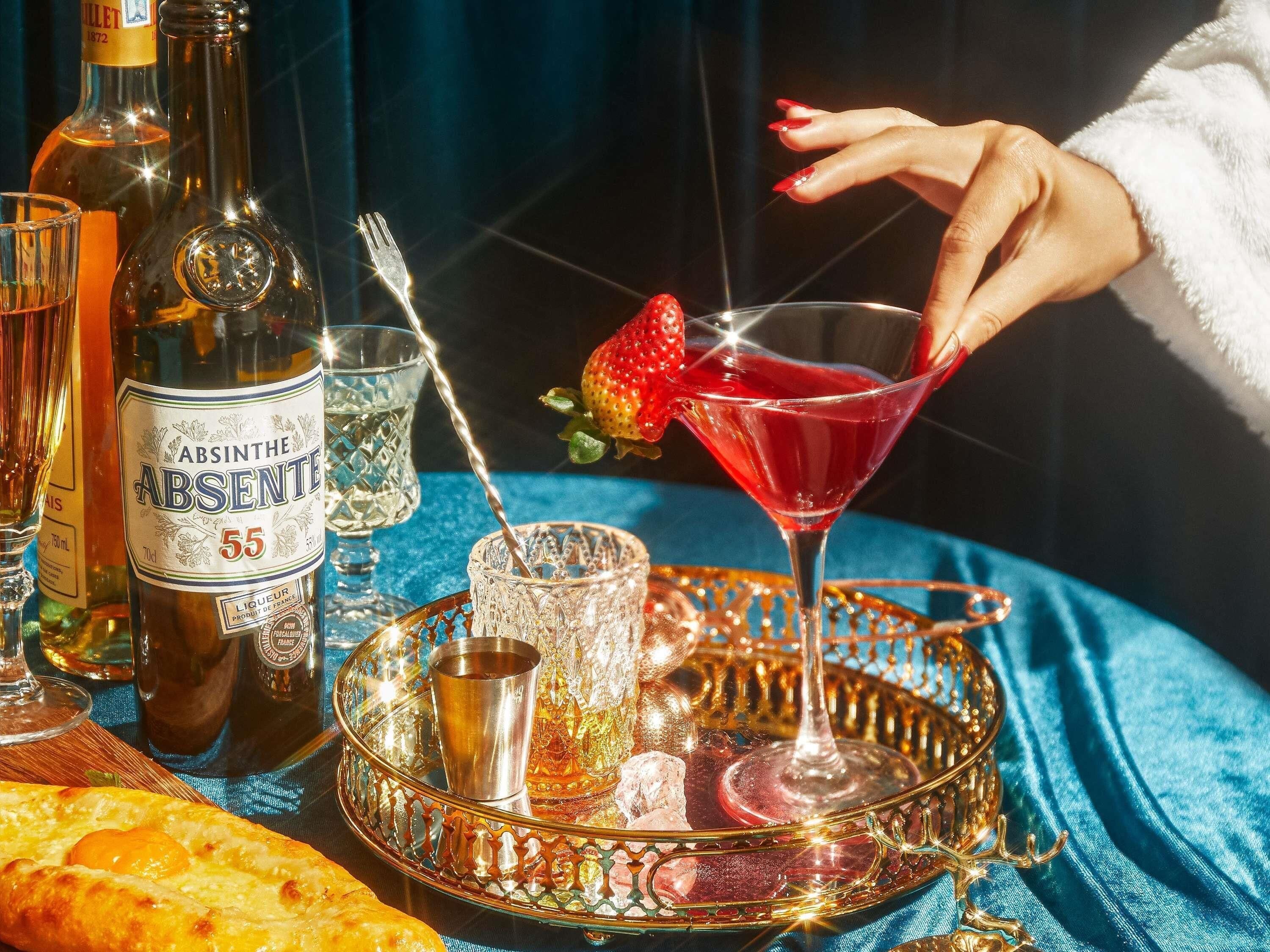 A tray with a martini glass, a bottle of absinthe, and a glass of red liquid with a strawberry garnish.