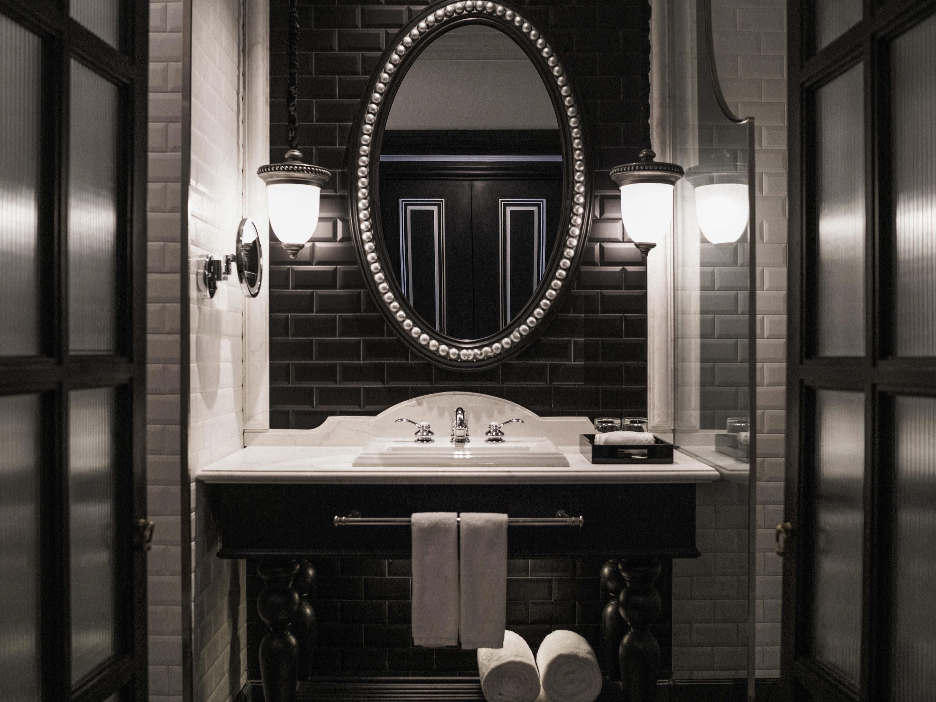 Un baño con pared de azulejos negros, un lavamanos blanco, un espejo grande con marco decorativo y dos luces empotradas.