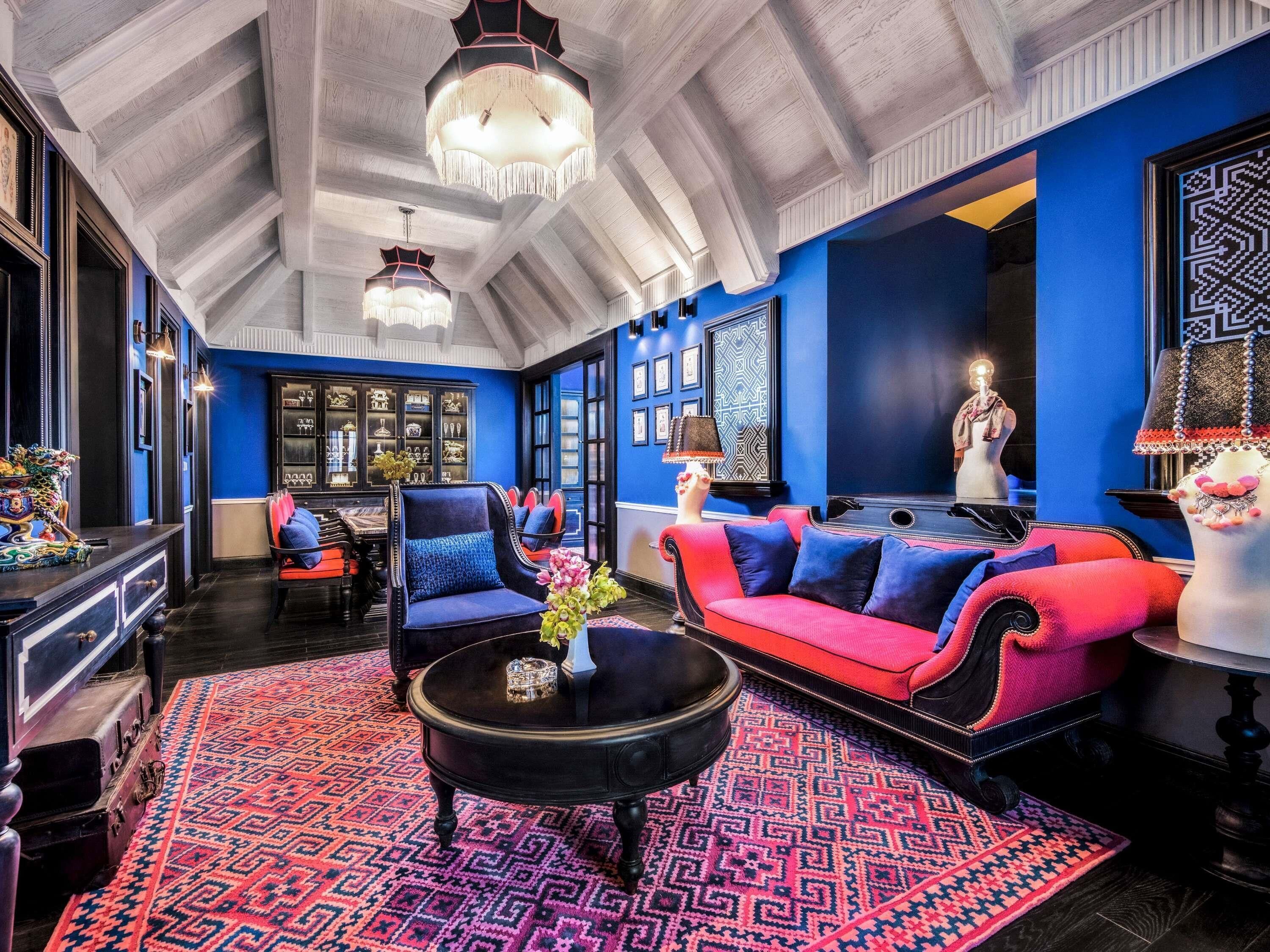 Una habitación con techo alto, alfombra estampada, sofá rojo y azul, mesa de centro negra y candelabro.
