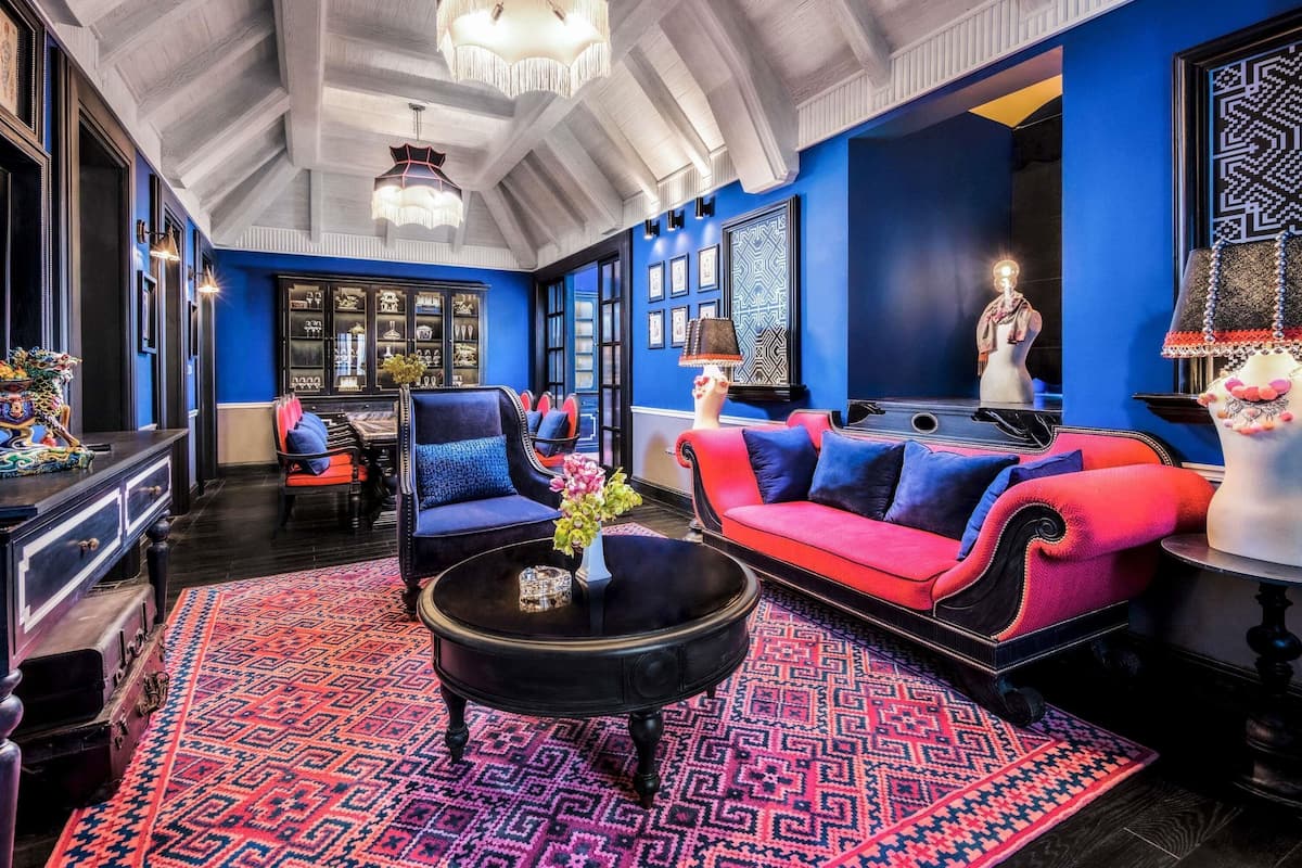 Una habitación con techo alto, alfombra estampada, sofá rojo y azul, mesa de centro negra y candelabro.