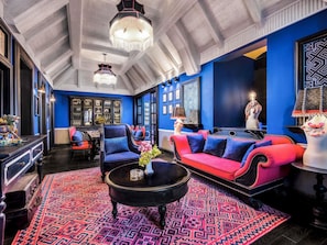 Una habitación con techo alto, alfombra estampada, sofá rojo y azul, mesa de centro negra y candelabro.