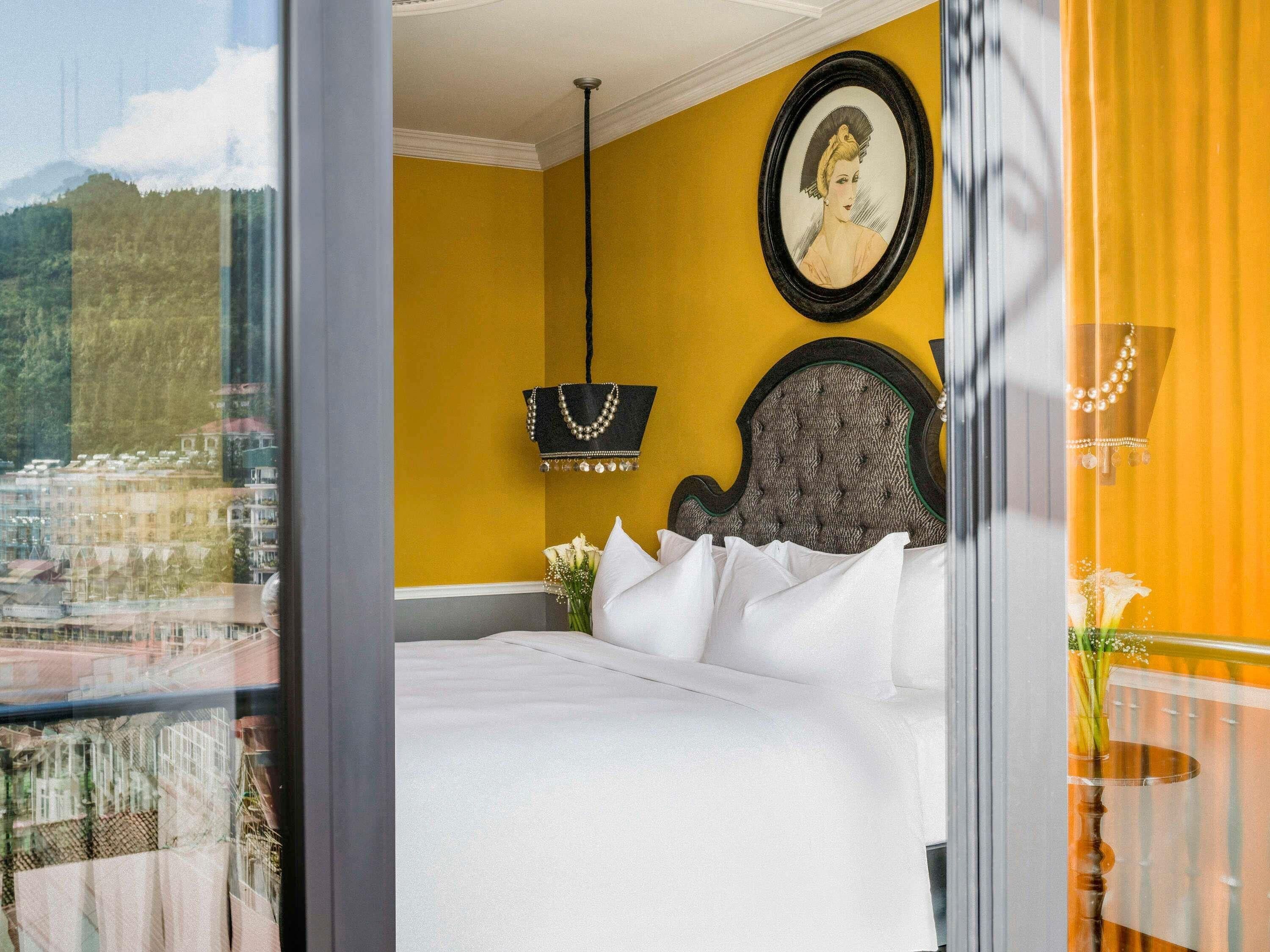 A hotel room with a bed, a framed portrait, a hanging handbag, and a view of a cityscape through a window.