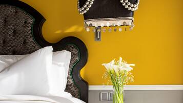 A bed with white linens, a round black table with a vase of white flowers, and a hanging black handbag with pearl accents against a yellow wall.