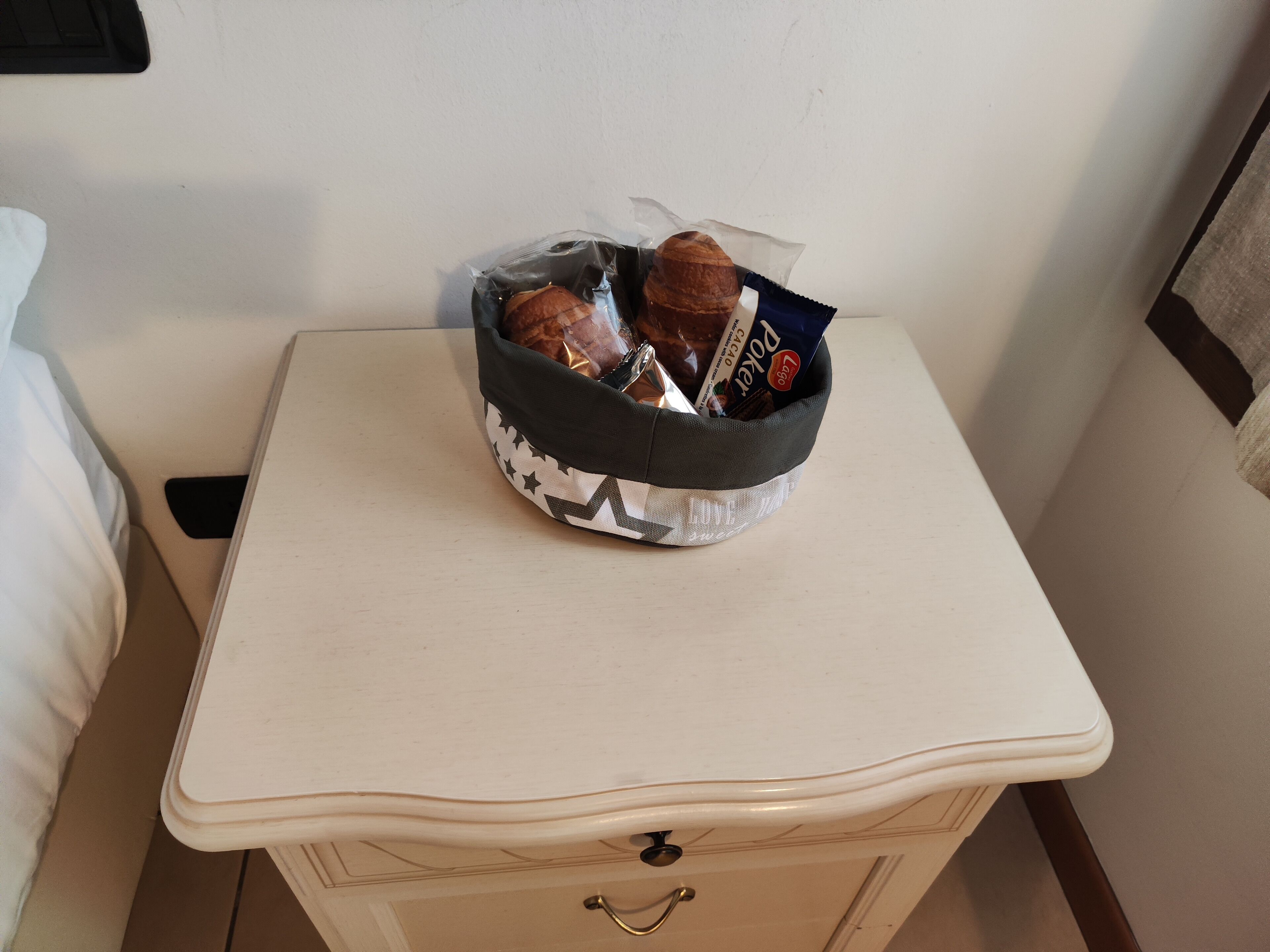 A small bedside table with a white drawer and a basket containing a croissant and a packet of coffee.