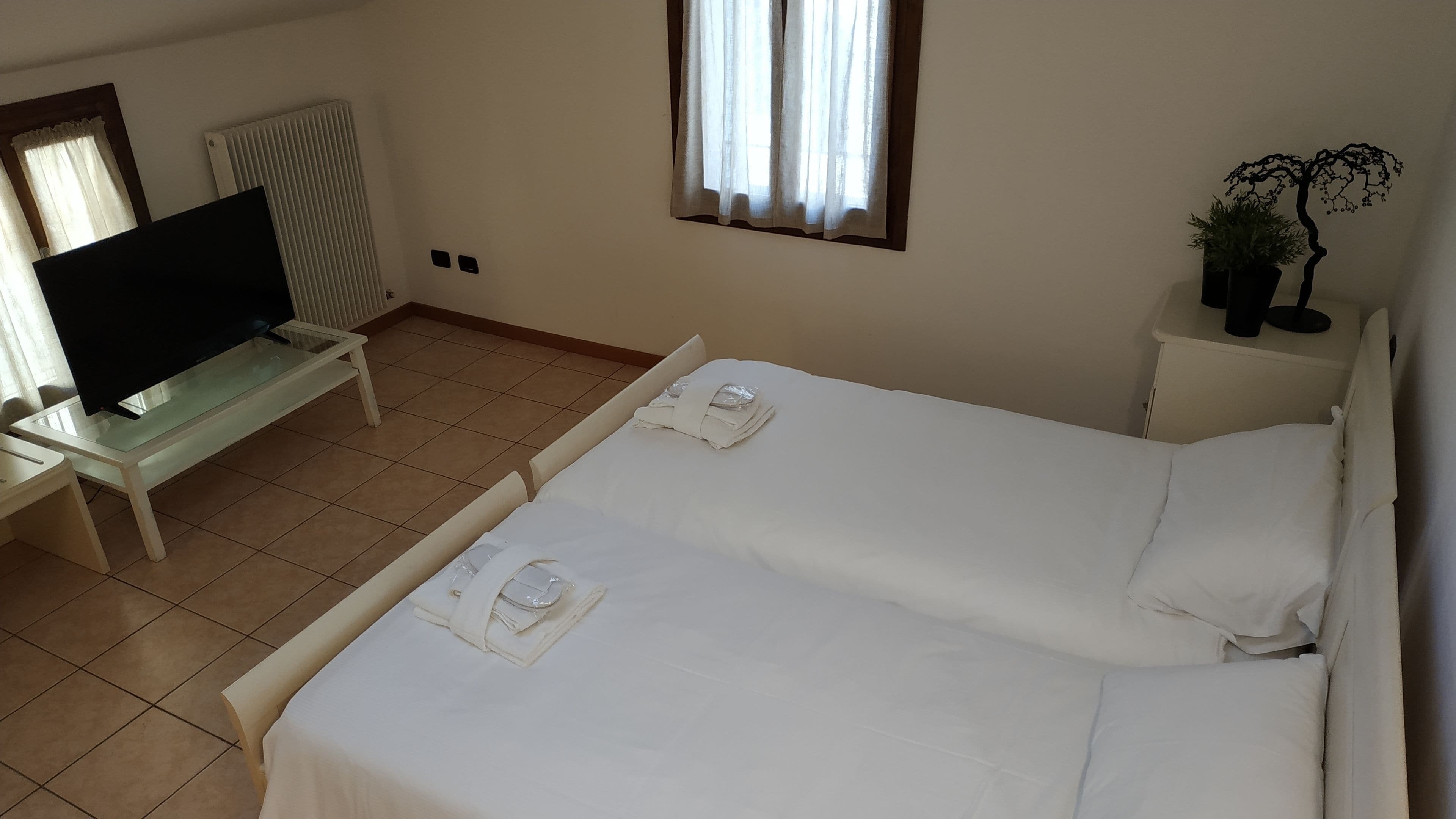 Une chambre d’hôtel avec un lit, un téléviseur posé sur une table en verre, un radiateur et une fenêtre avec des rideaux.