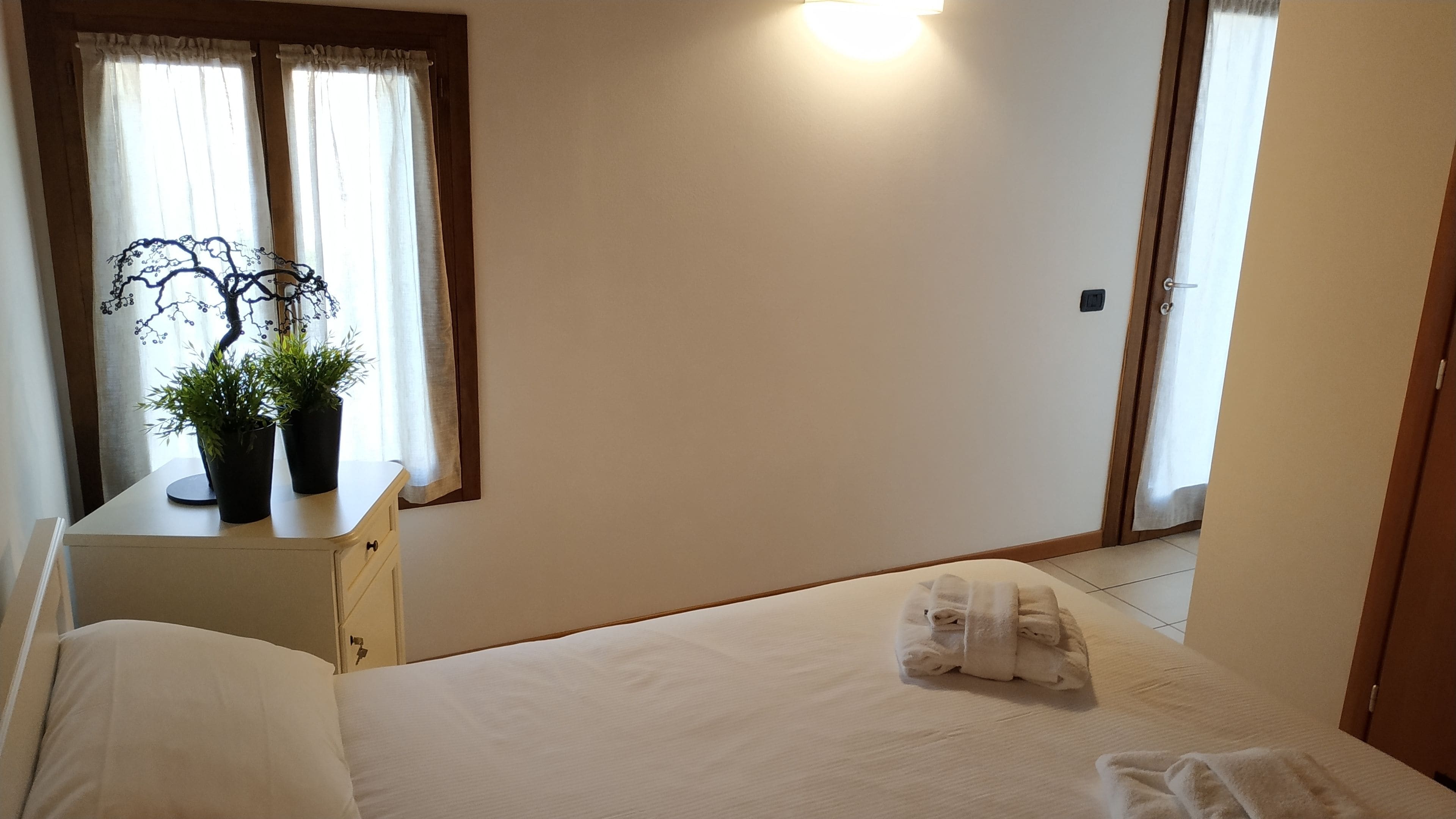 Un dormitorio con cama, una mesita de noche con dos plantas en macetas, una ventana con cortinas y una puerta.