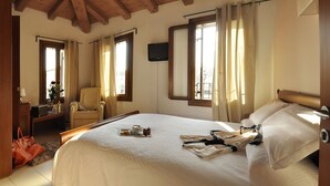 Quarto de hotel com uma cama grande, uma cadeira, escrivaninha com abajur e televisão na parede.