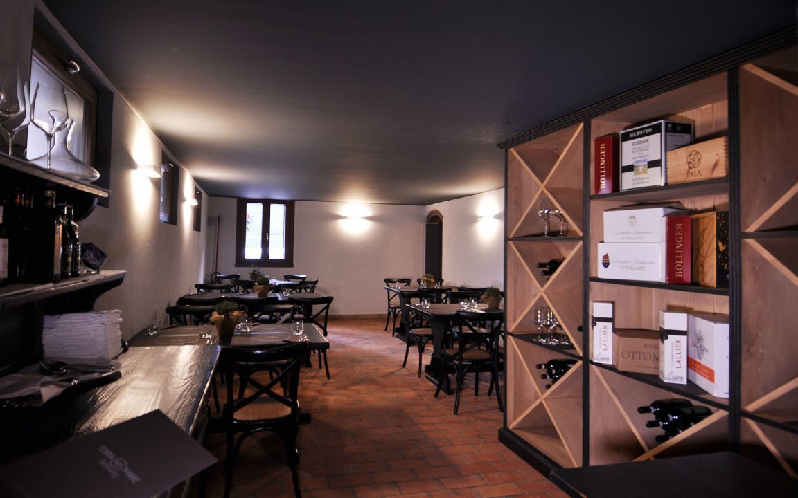 Een restaurant met meerdere tafels en stoelen, een wijnrek vol flessen, en een vloer van baksteen.
