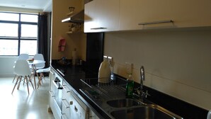 Una cocina moderna con armarios blancos, encimera negra y pileta de acero inoxidable. Se ve una ventana que proporciona luz natural y una zona de comedor con sillas blancas.