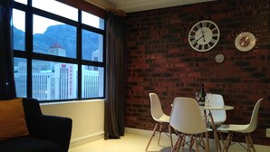 Una habitación con una mesa de comedor, sillas, un sofá, un ventanal con vista a un edificio y una pared de ladrillo con relojes.