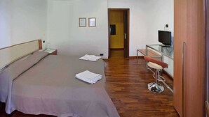 Quarto de hotel com cama, escrivaninha com cadeira, televisão e chão de madeira.