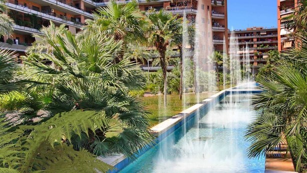 Um hotel com piscina, apresentando uma fonte de água, rodeado por palmeiras e vegetação.
