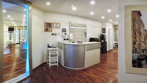 Uma área de recepção de hotel com balcão, placa com 'LA FIERA BED & BREAKFAST', telefone, dispensador de água e uma moldura com imagem na parede.