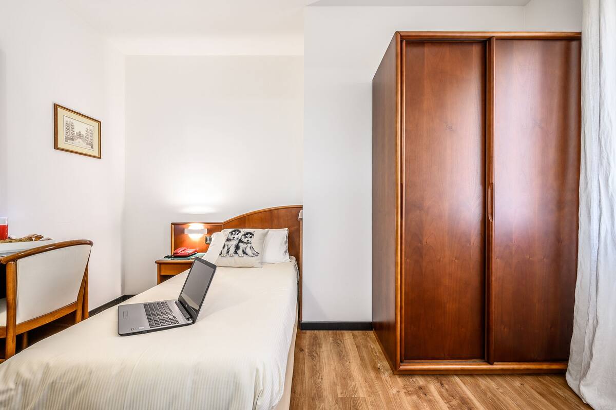 Ein Hotelzimmer mit einem Bett, einem Laptop, einem Holzschrank, einem Schreibtisch mit Stuhl und einem gerahmten Bild an der Wand.