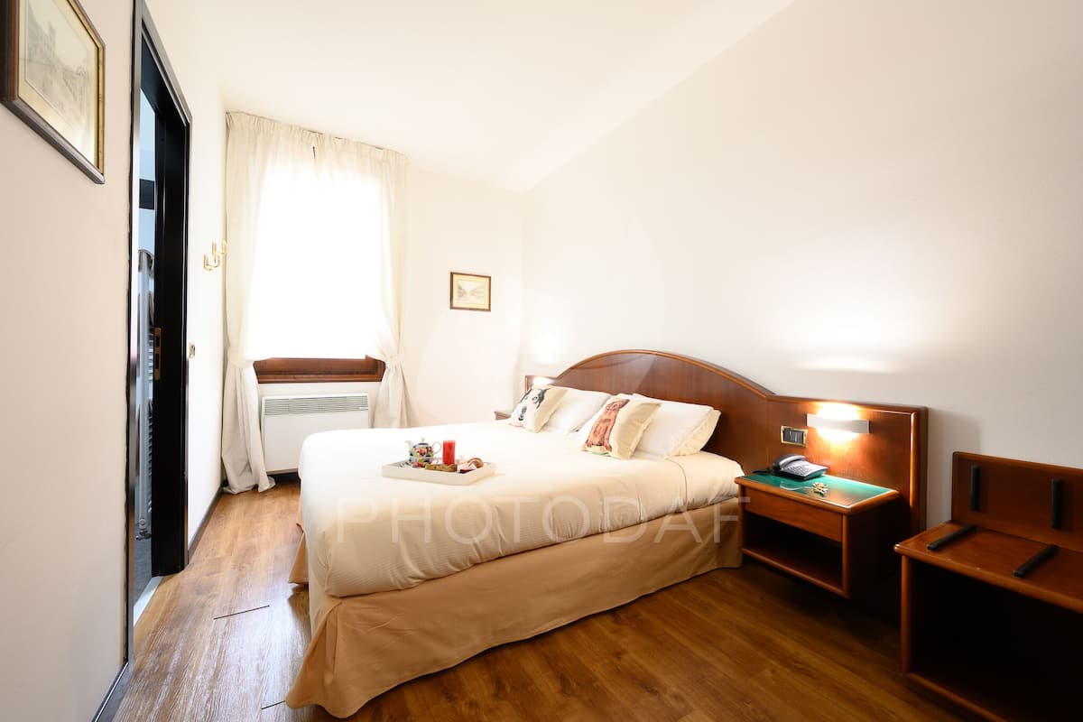 Ein Hotelzimmer mit einem Holzbett, einem Nachttisch mit Lampe, einem Fenster mit Vorhängen und einem gerahmten Bild an der Wand.