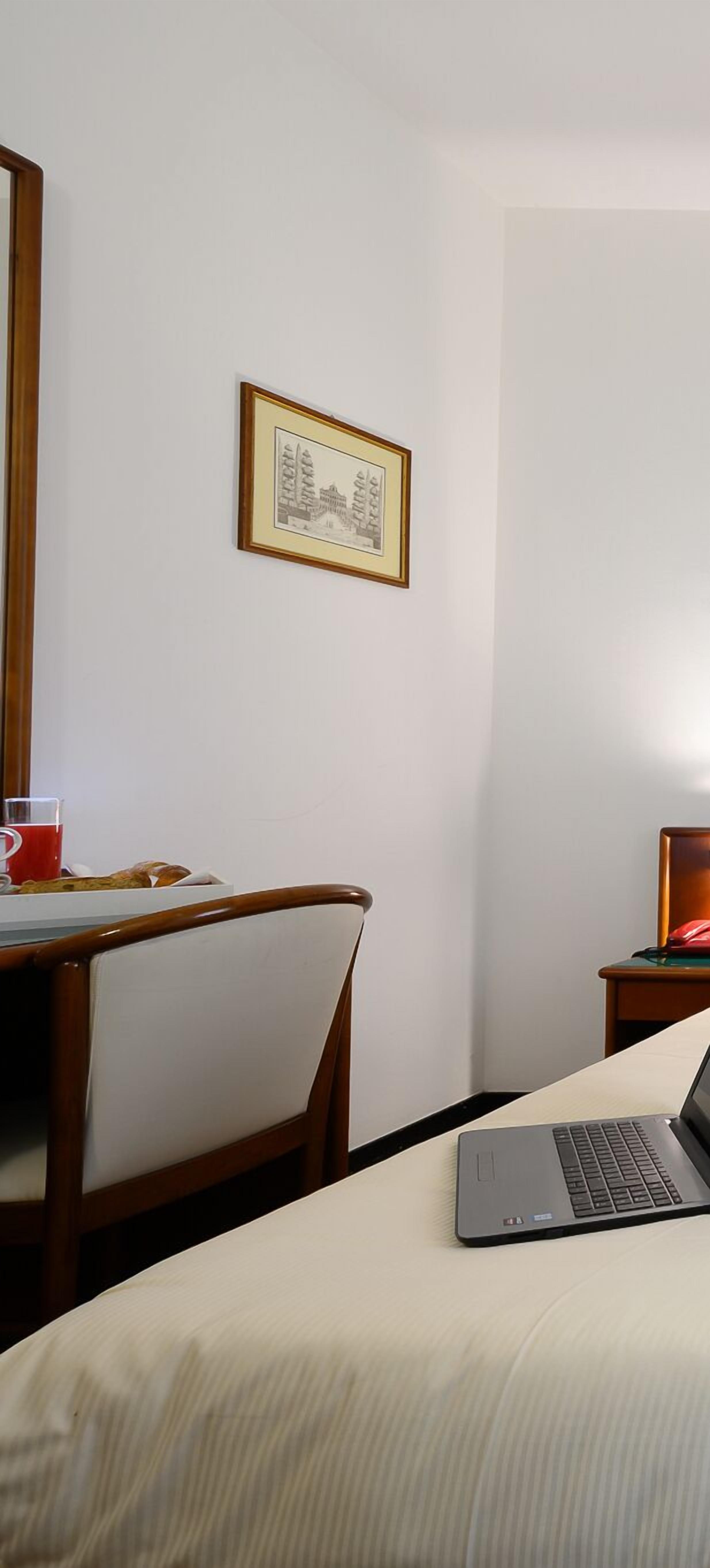 Hotelkamer met een bed, een laptop op het bed, een stoel, een kleine tafel met een rode beker en een glas, en een ingelijst foto aan de muur.