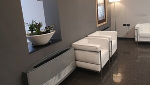 Un área de espera moderna con una planta grande en maceta, un extintor en la pared, un cuadro enmarcado y una lámpara de pie.