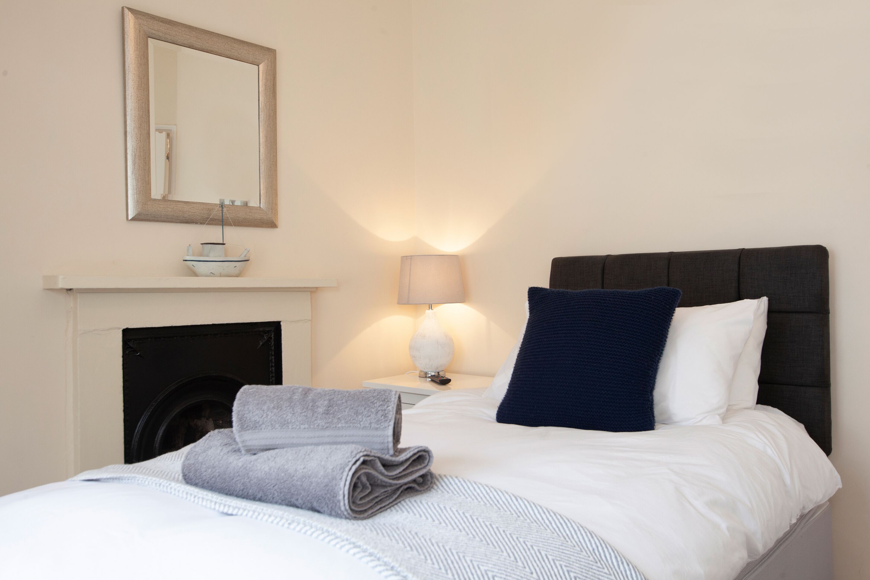 En pænt redt seng med et hvidt og gråt stribet tæppe, et gråt håndklæde lagt sammen, en mørkeblå pude og en hvid pude. Der er et lille hvidt bord med en lampe og en fjernbetjening ved siden af sengen. En pejs er synlig i baggrunden med en dekorativ genstand ovenpå.