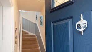 Eine Treppe mit beigem Teppich führt zu einer Zwischenebene, dort befindet sich eine blaue Tür mit silberner Klopffunktion und ein kleines Fenster mit geometrischem Muster.