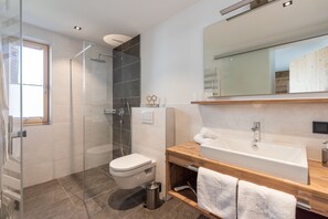 A modern bathroom with a glass shower enclosure, a white toilet, a wooden vanity with a sink, and a large mirror. - Fetz Luxury Appartements (Steeg)