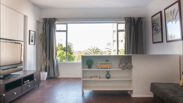 Una sala de estar moderna con un televisor de pantalla plana, una estantería con objetos decorativos y un amplio ventanal con cortinas.