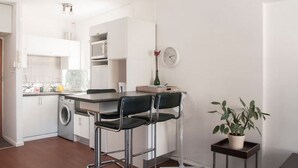 Cocina compacta con lavadora, microondas y un pequeño comedor con dos sillas y un ventilador.