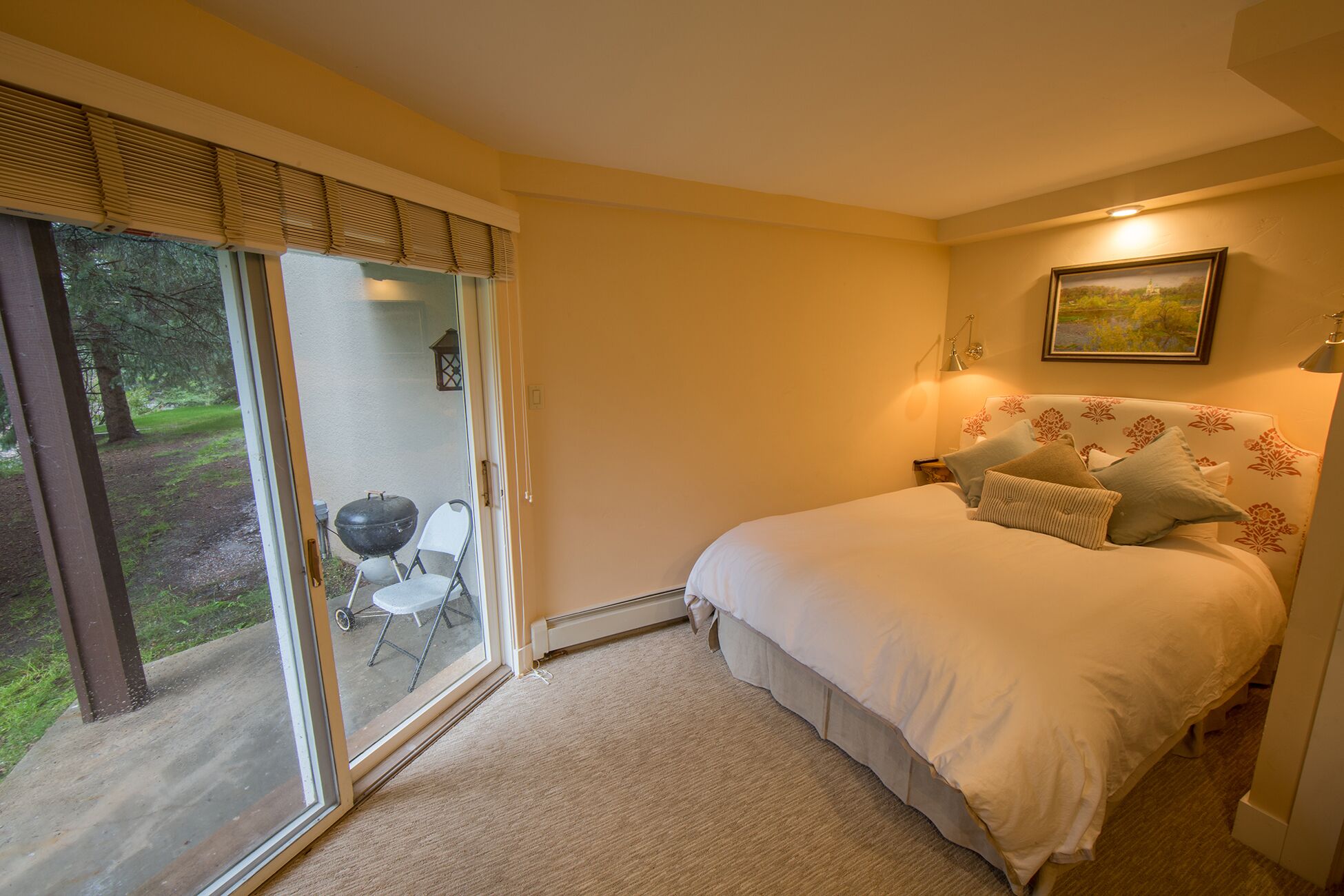 A bedroom with a bed, a nightstand, a lamp, a painting, and a sliding glass door leading to an outdoor area with a barbecue grill and chairs.
