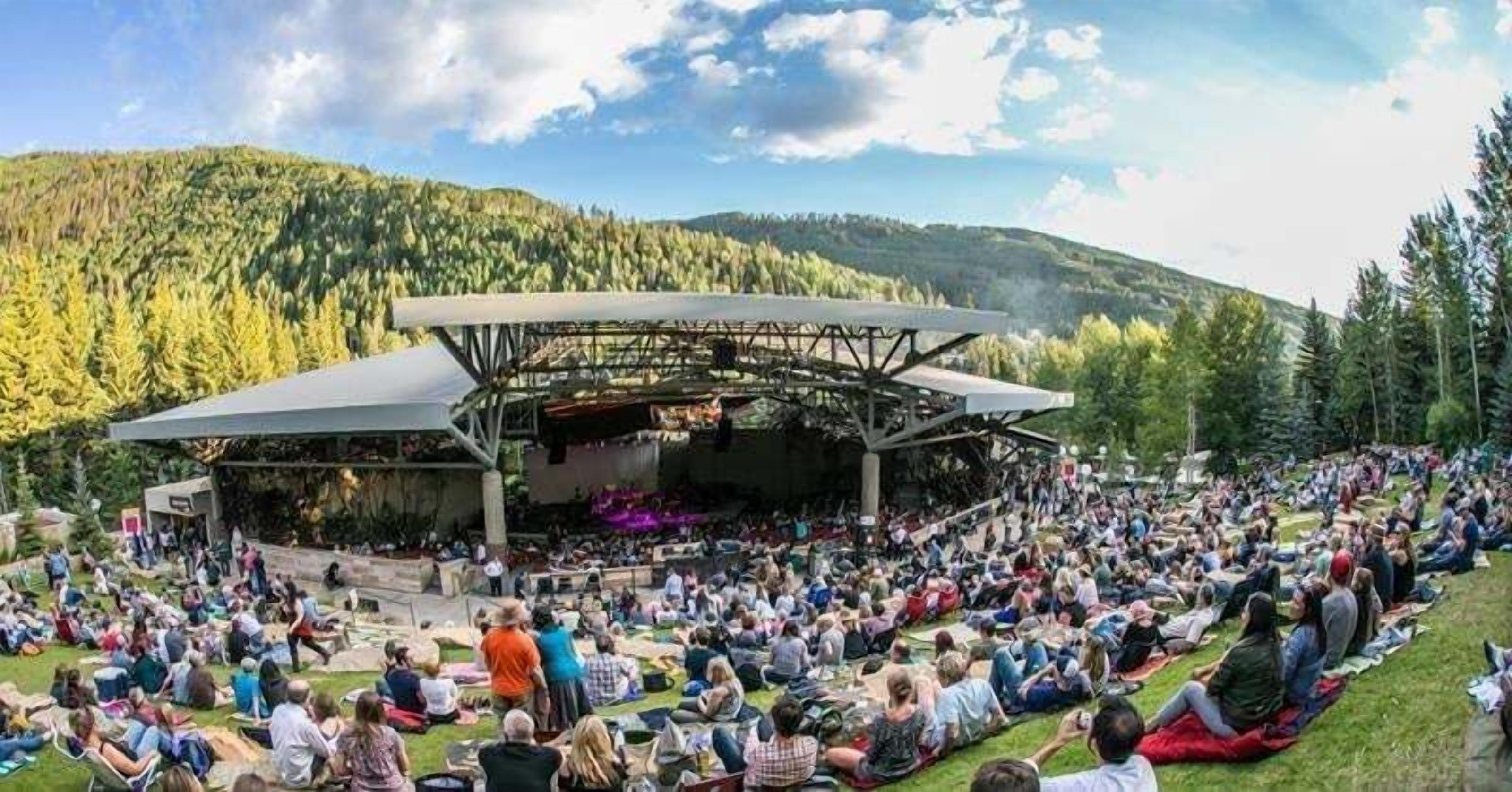 An outdoor amphitheater with a stage, surrounded by a grassy area with people seated.