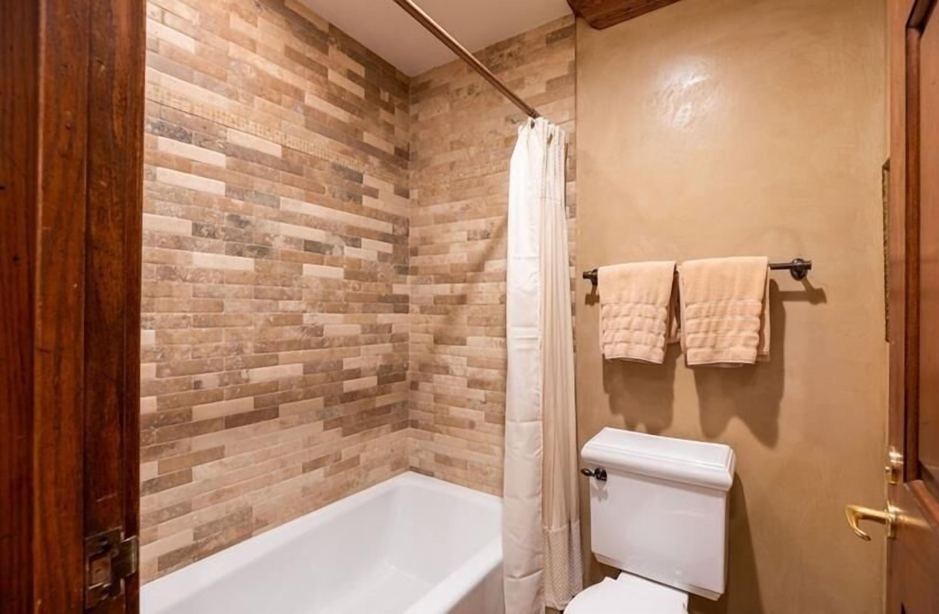 A bathroom with a brick-patterned wall, a white bathtub, a white toilet, and two towels hanging on a towel rack.