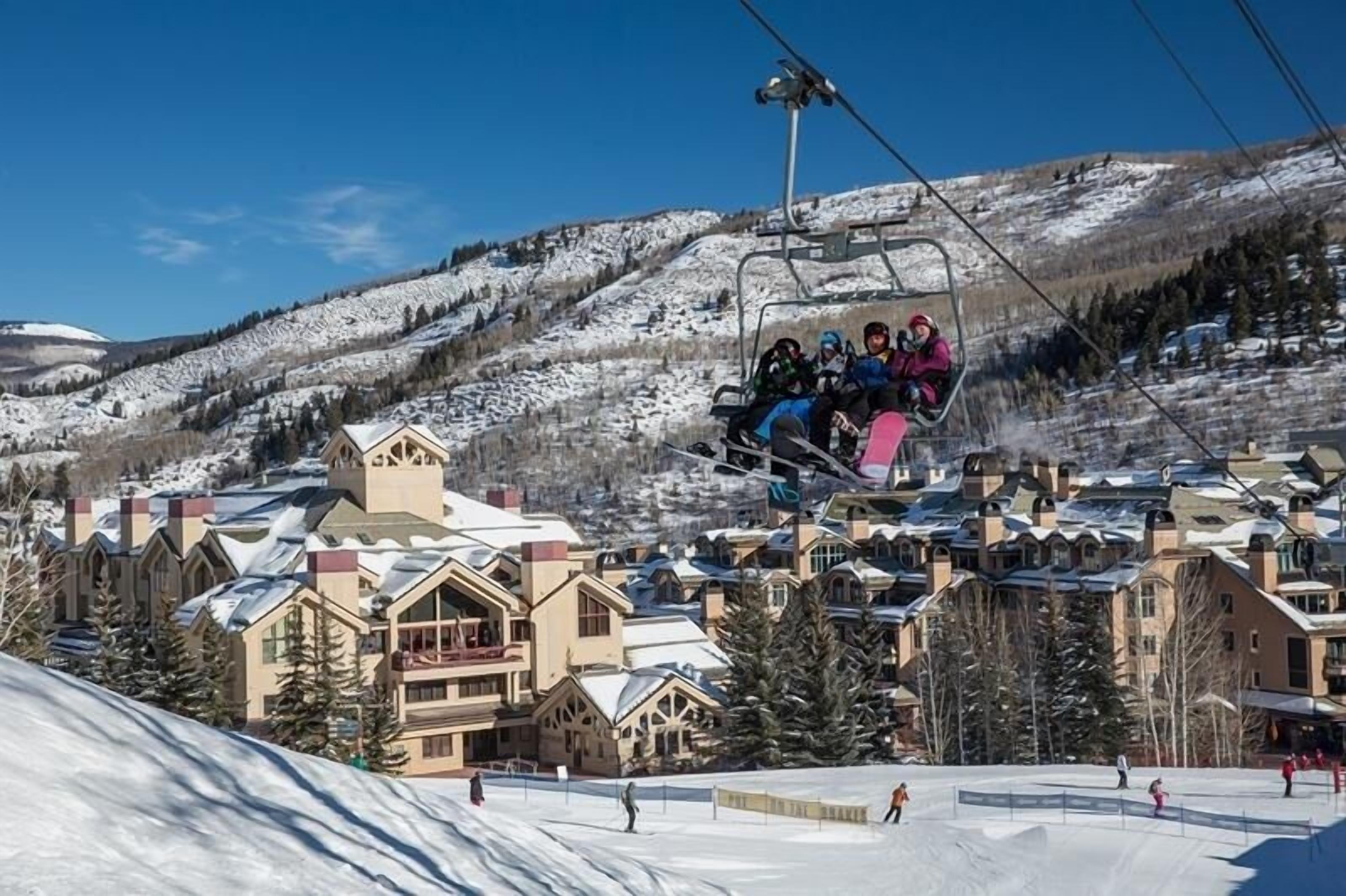 A ski lift with people seated, a snow-covered mountain, and buildings with a traditional architectural style.