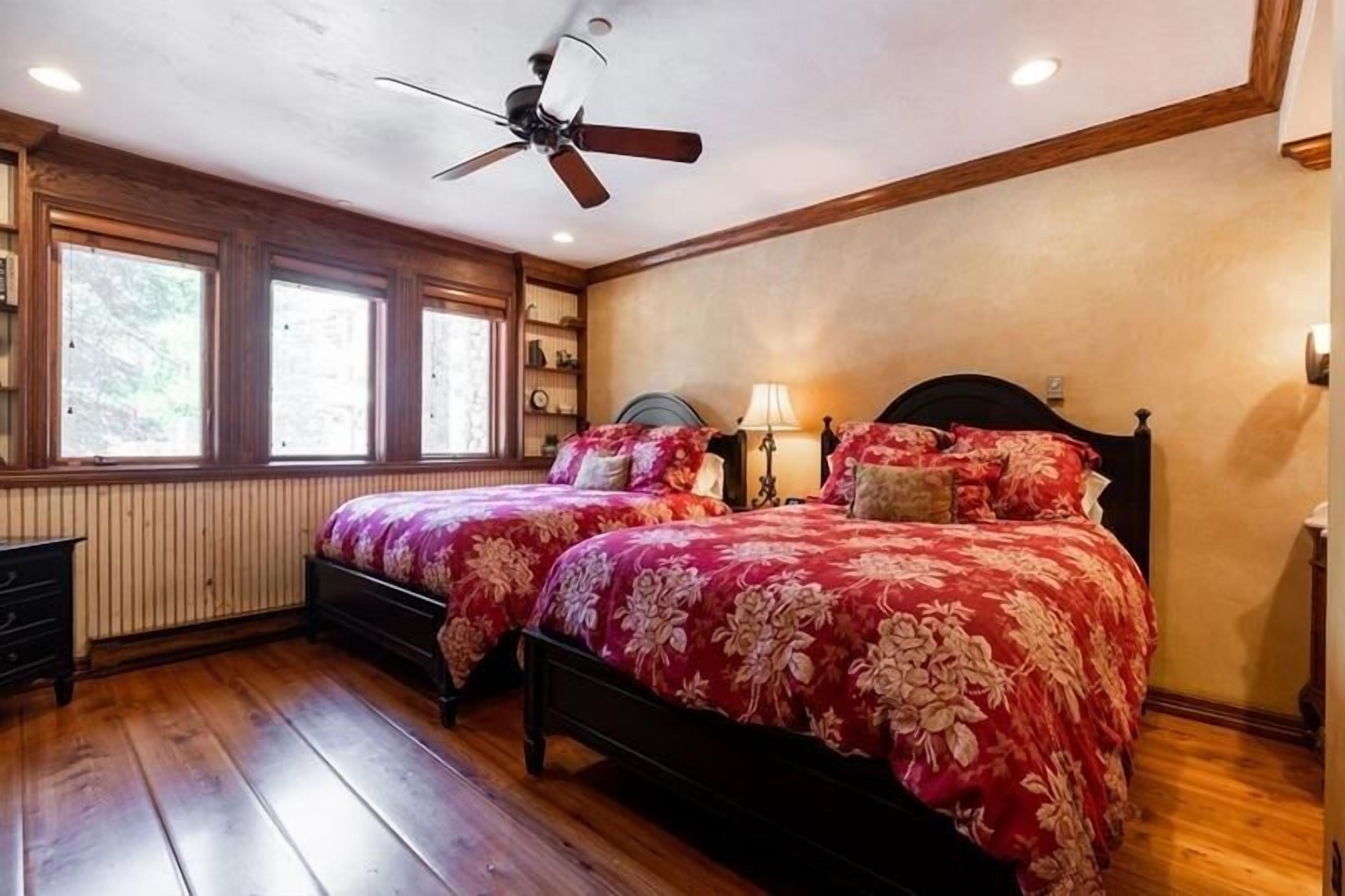 A bedroom with two beds, wooden flooring, a ceiling fan, and large windows.