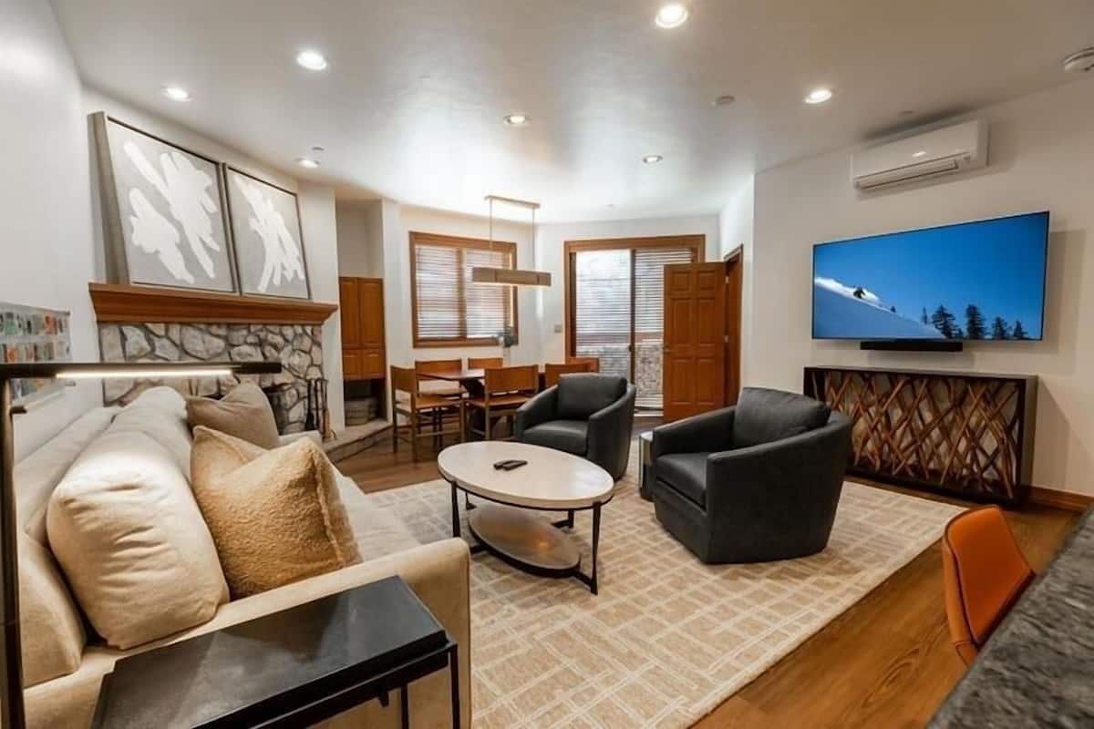 Een moderne woonkamer met een bank, fauteuil en salontafel. Er is een televisie aan de muur bevestigd en een open haard met een stenen omlijsting.