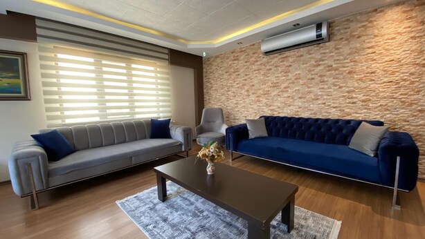 Una sala de estar moderna con un sofá gris, un sofá azul, un sillón gris y una mesa de centro. Hay una alfombra estampada en el suelo, y un equipo de aire acondicionado está montado en la pared.