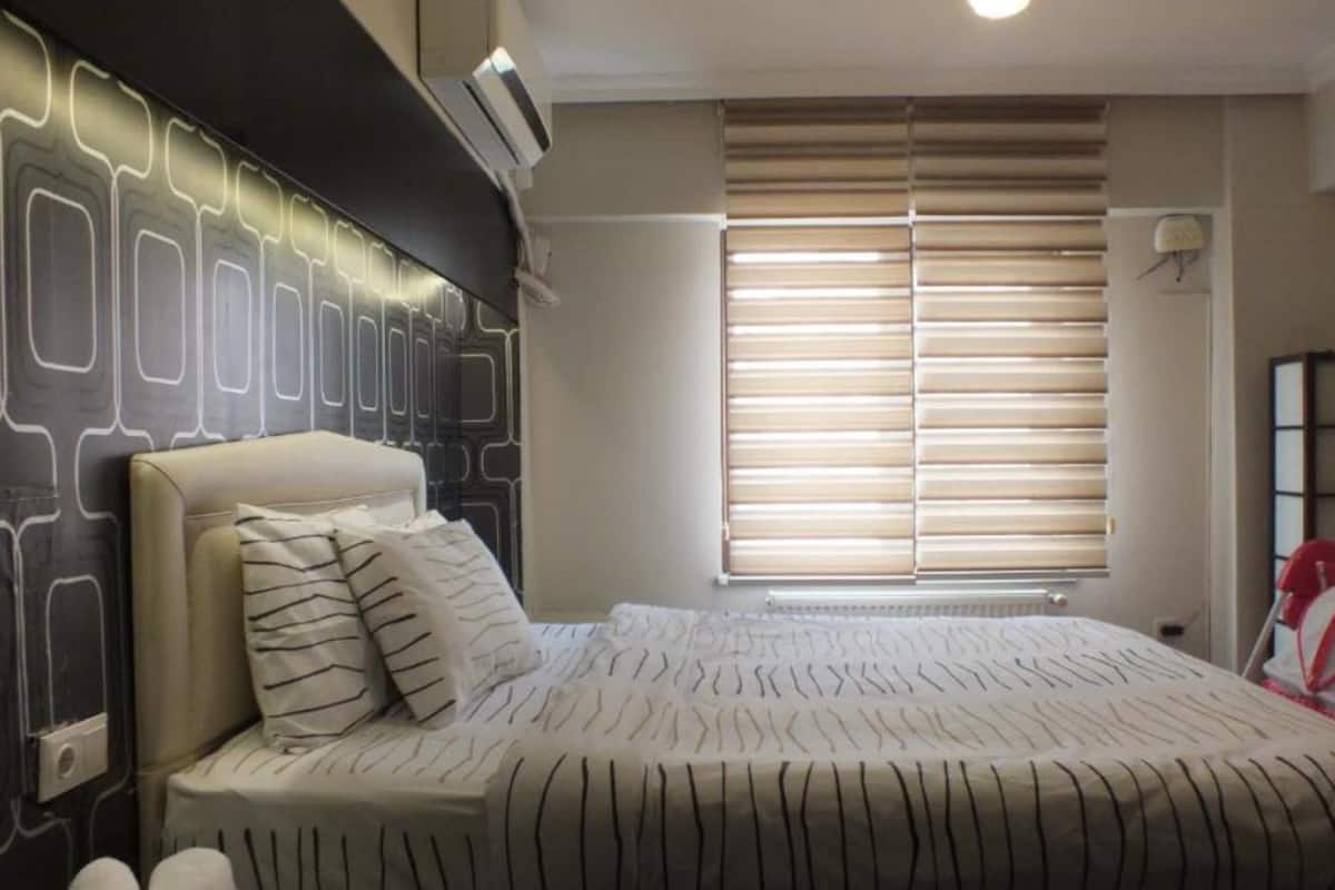 A bedroom with a bed, a headboard, a window with blinds, and a wall-mounted air conditioner.