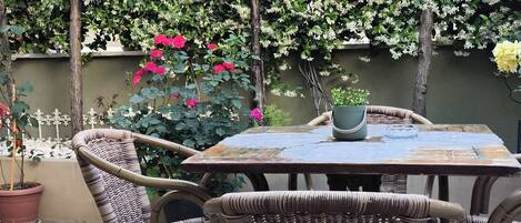 An outdoor patio with a round table and wicker chairs, surrounded by potted plants and flowering shrubs.
