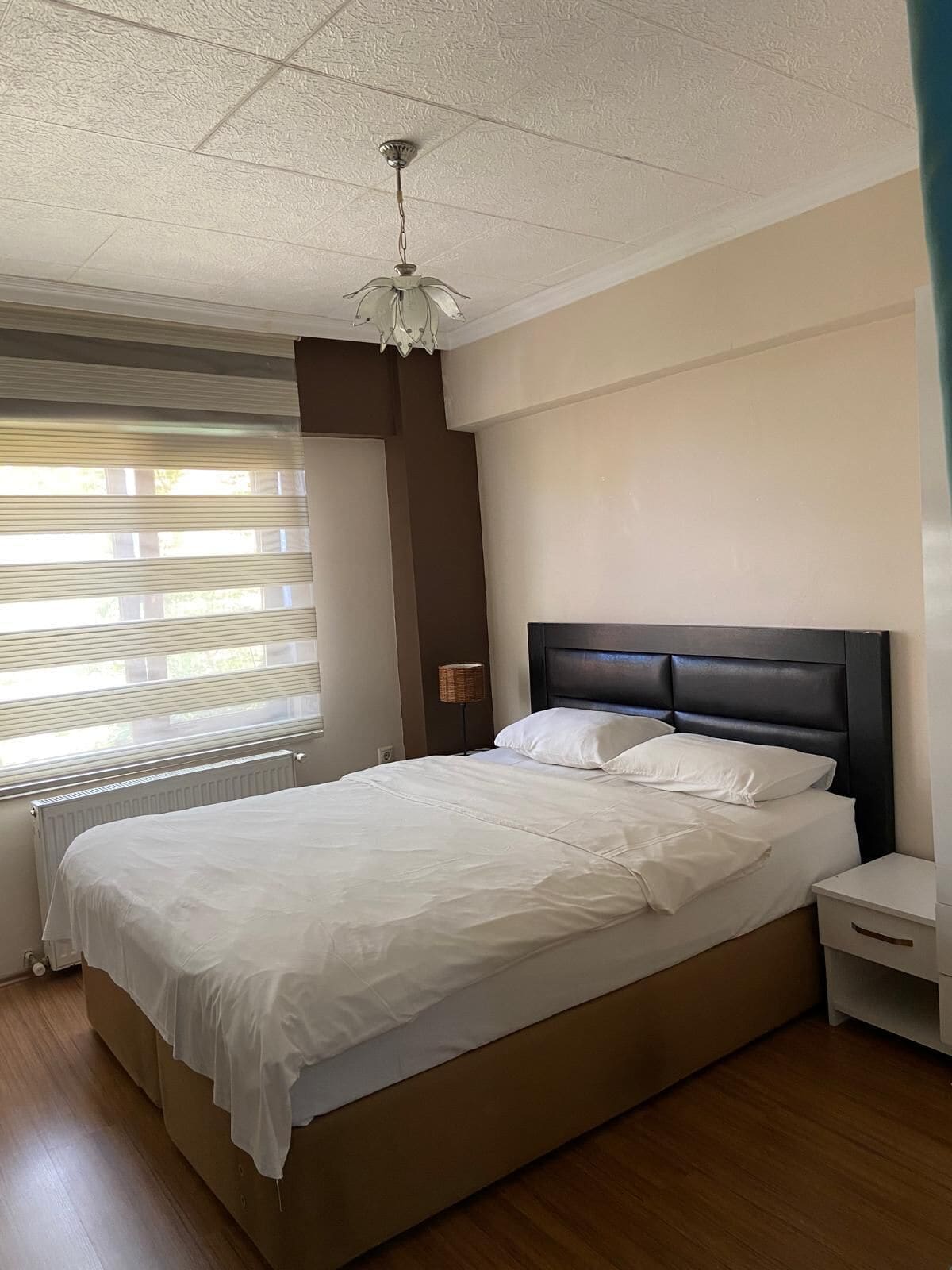Una cama bien hecha con sábanas blancas en una habitación con suelo de madera, cabecero oscuro y ventilador de techo.